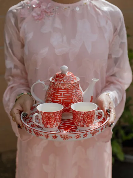 Tea ceremony set with red double happiness teapot and cups on a tray, held by a bride in a pink embroidered qipao in a garden setting