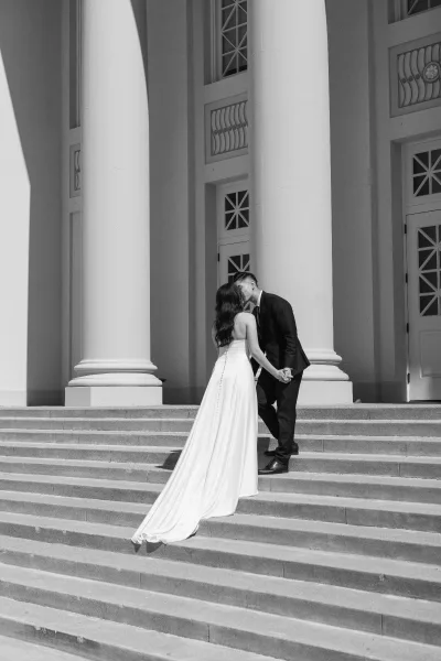 Wedding kiss portrait of bride and groom kissing, holding hands on stone steps before white columns, her satin gown with long train