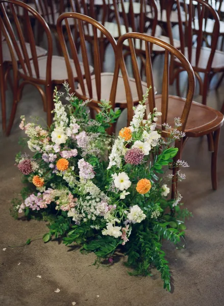 Ceremony aisle flowers in a meadow arrangement of greenery with orange and pastel blooms lining wooden chairs on a concrete floor indoors