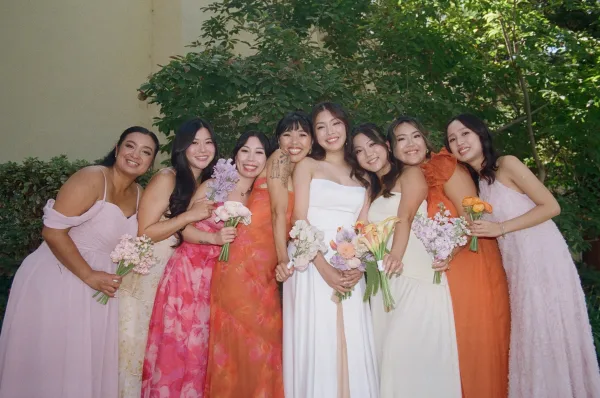 Bridesmaids group photo with bride and bridesmaids holding bouquets in colorful mismatched dresses, posed outdoors by trees and a stucco wall