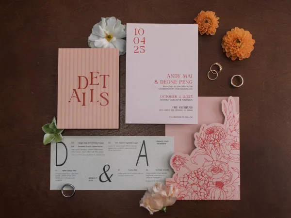 Invitation suite flat lay with pink envelope, floral-printed cards, and gold and silver rings beside orange flowers on a wood tabletop