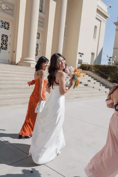 Bride portrait holding bouquet, looking back in a strapless white dress with bridesmaids in orange and pink on sunny courthouse steps