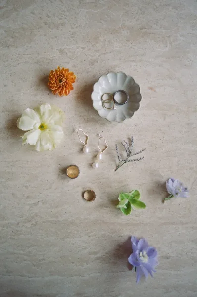 Wedding rings flatlay with engagement ring and wedding band set on a ceramic dish beside pearl drop earrings and floral greenery on stone surface