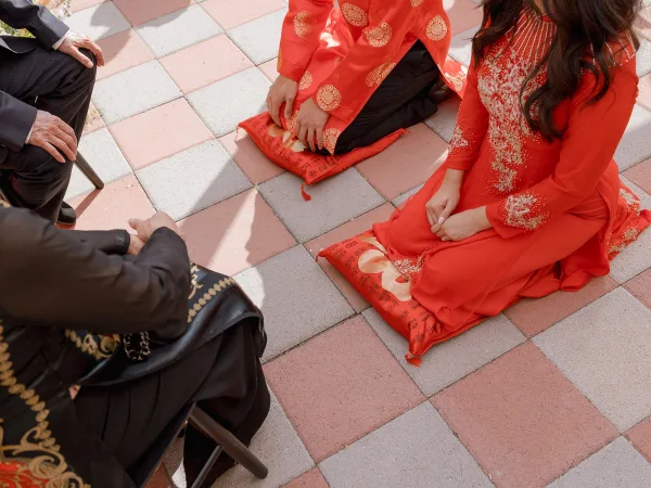 Tea ceremony with couple kneeling on cushions in red embroidered attire, serving tea to seated parents on a sunlit patio