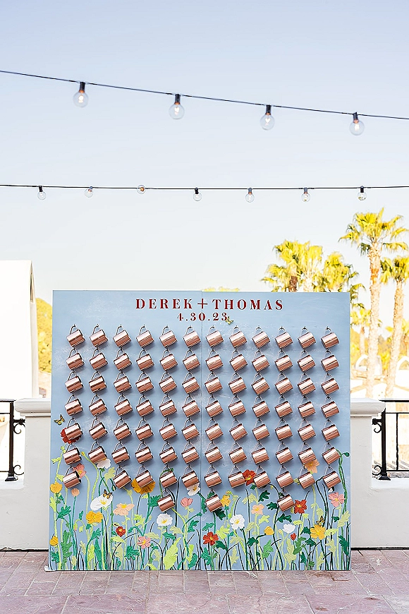 Wedding seating chart escort card wall with copper mugs and painted floral illustration, lit by string lights on an outdoor terrace with palms