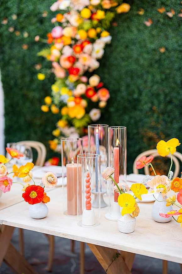 Reception tablescape with colorful wedding centerpieces in bud vases and taper candles in glass hurricanes on a white table, hedge wall patio backdrop