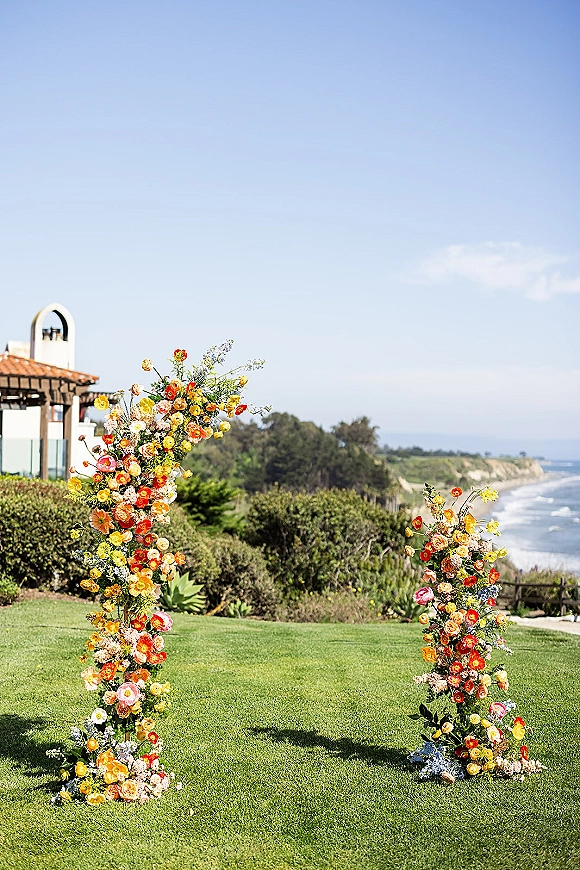 Wedding ceremony arch with an asymmetrical floral arch of colorful roses and ranunculus on a lawn overlooking the ocean coastline under open sky