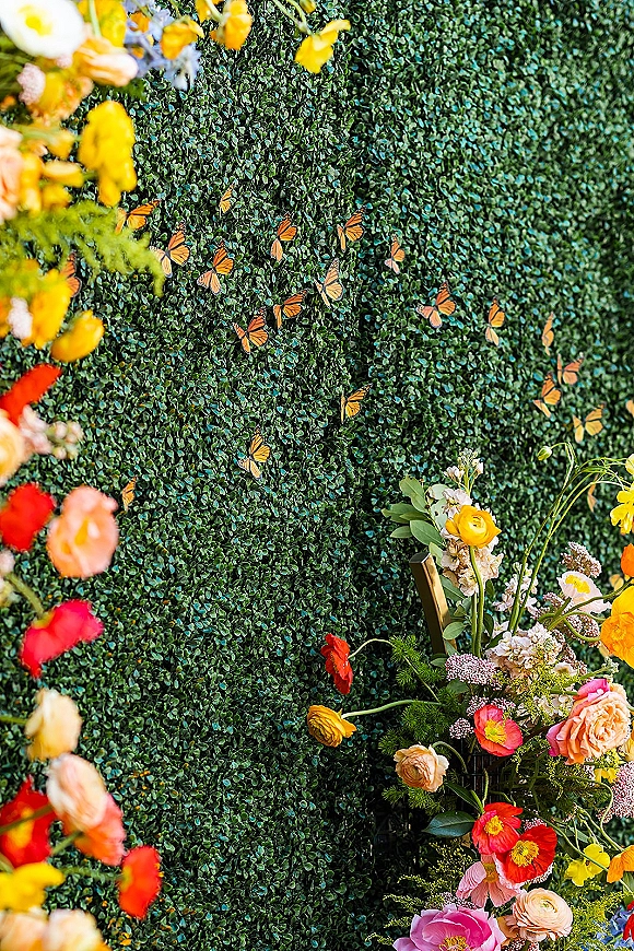 Wedding photo backdrop with boxwood hedge backdrop, bright roses and wildflower blooms in a brass frame with butterfly accents