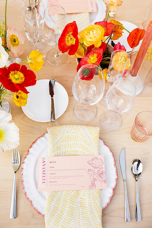 Wedding tablescape with colorful wedding tablescape poppy and ranunculus centerpiece, taper candles, place cards, and mismatched glassware on light wood table