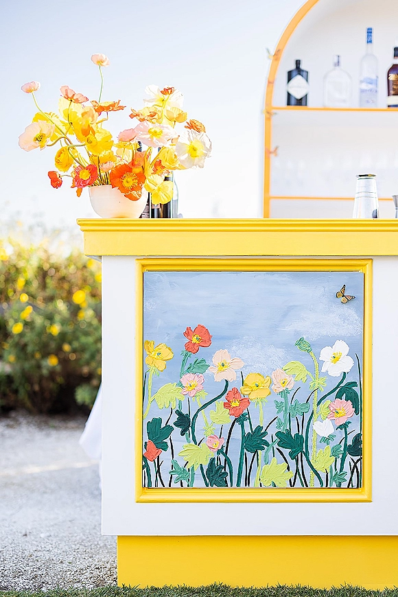 Wedding bar setup with a bright yellow counter, liquor bottles and glassware, plus a painted floral panel in an outdoor garden setting