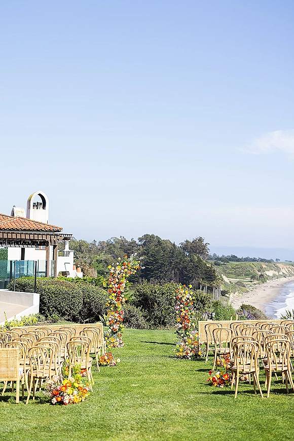 Ceremony setup with a floral arch and aisle florals, rattan chairs on a lawn facing the ocean and cliffs near a villa
