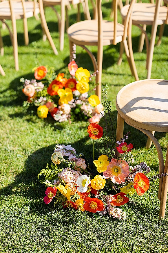 Ceremony aisle flowers in grounded clusters of poppy and ranunculus with peach, yellow, and orange blooms beside wooden chairs on a grass lawn