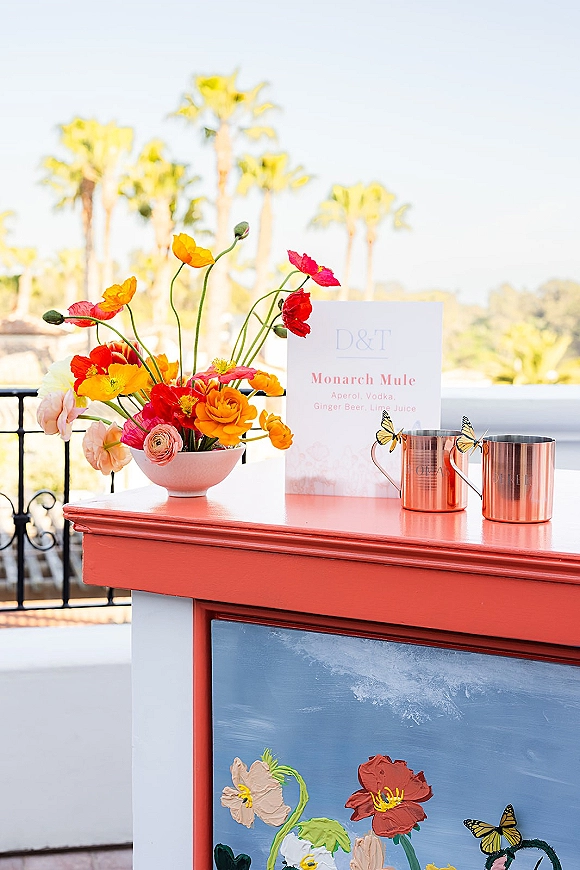 Signature cocktail display with wedding signature drink sign, copper mugs and butterfly stirrers beside florals on an outdoor terrace with palms