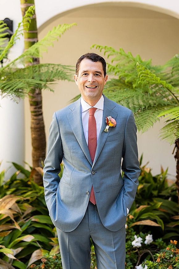 Groom portrait of a smiling man in a gray suit with coral tie and boutonniere, hands in pockets by a tropical courtyard archway