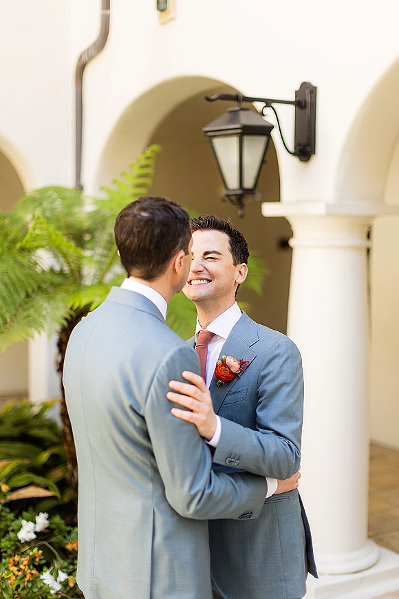 Groom portrait of two grooms in light blue suits with boutonnieres embracing under a white arched colonnade in a lush courtyard