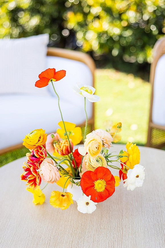 Wedding centerpiece with a bright floral arrangement of poppy and ranunculus in a small vase on a garden table with wooden chairs