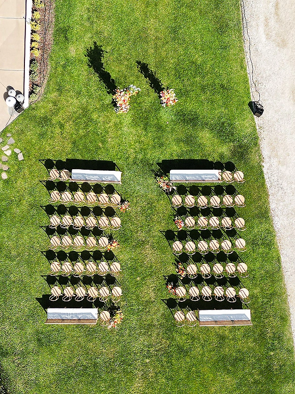Ceremony seating layout with two sections of wood chairs lining a white aisle runner, floral arrangements and benches on a lawn walkway