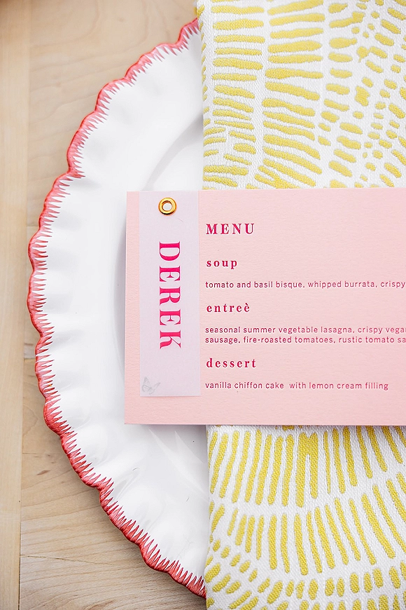 Wedding menu card in pink with a menu tag and grommet, styled on a scalloped edge plate with patterned napkin on a light wood table