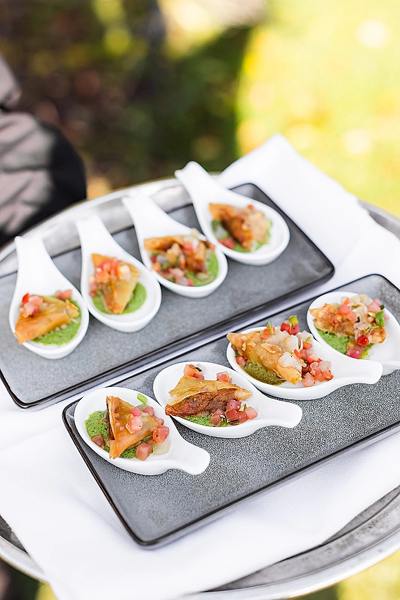 Wedding appetizers served on passed appetizer spoons, neatly arranged on black plates with white ceramic spoons on a tray outdoors