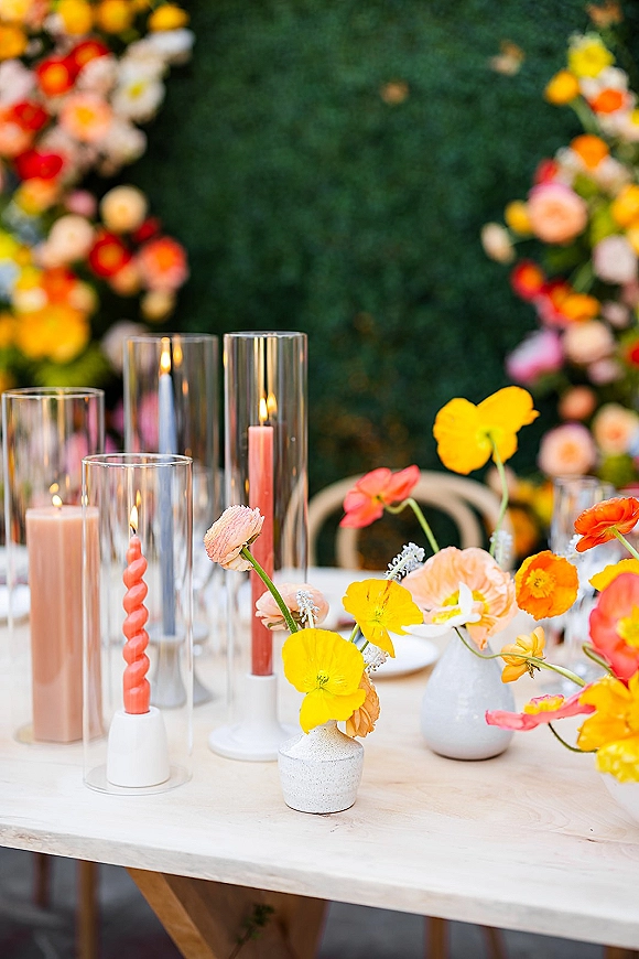 Reception tablescape with colorful wedding centerpieces in bud vases, pillar and spiral taper candles on a white table before a green hedge wall