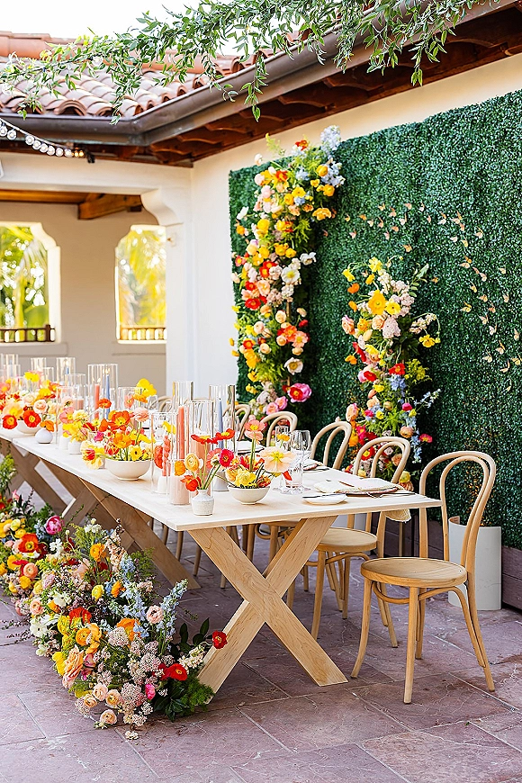 Reception tablescape with long head table decor, colorful bud vases and white taper candles, set in an outdoor courtyard with stucco arches