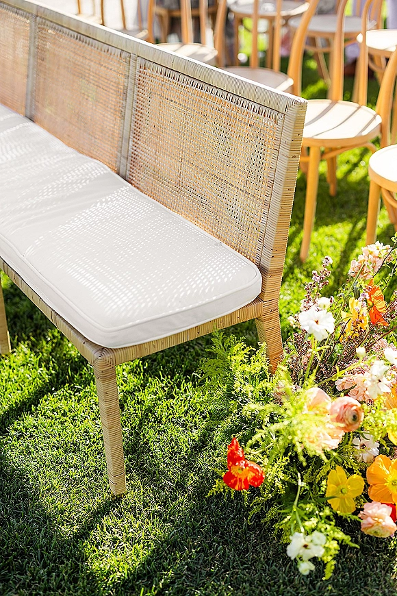 Ceremony seating with a rattan wedding bench and white cushion beside wooden chairs, accented by bright wildflowers on a sunlit lawn