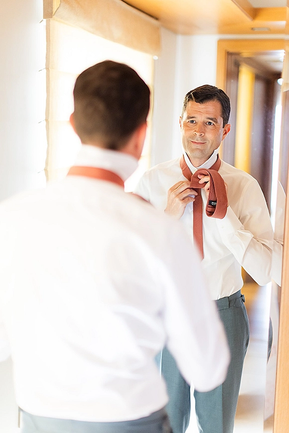 Groom getting ready, tying a rust tie over a white dress shirt in a mirror-lit hallway with string lights and a door frame behind