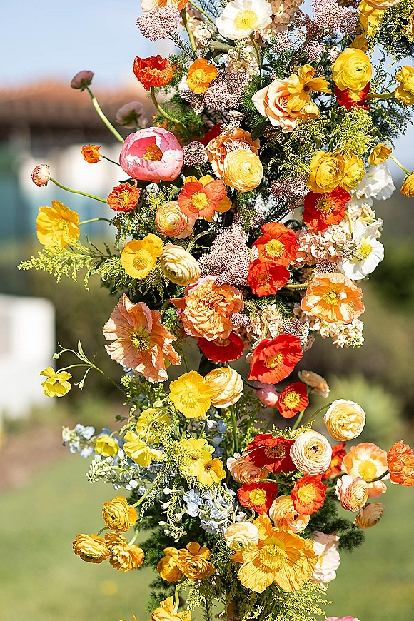 Wedding floral installation with ceremony arch flowers in pink, orange, and yellow blooms with greenery on an outdoor lawn with trees and sky