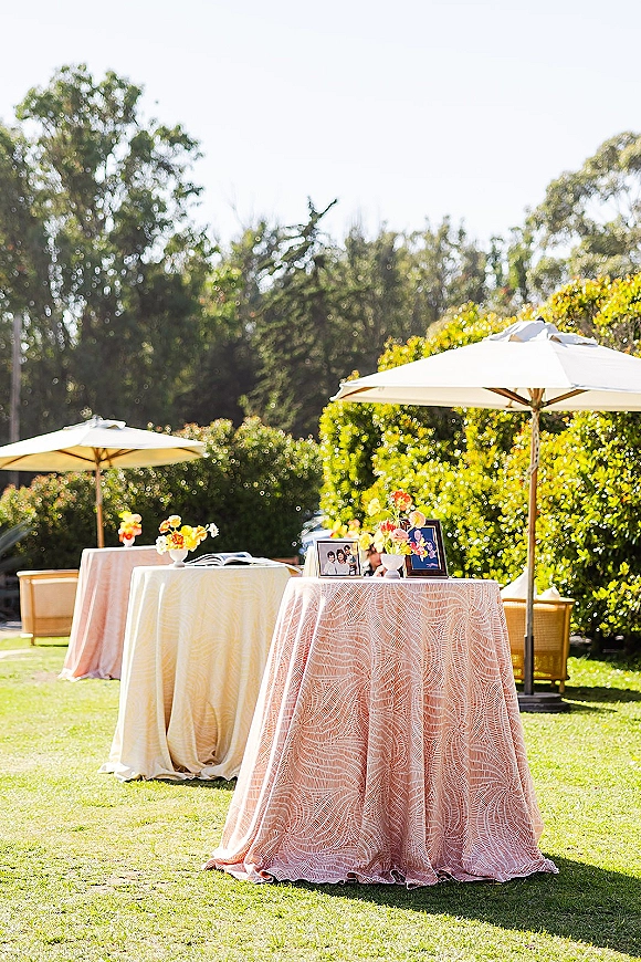 Cocktail table decor with outdoor cocktail tables draped in blush linens and lace overlays, bud vases, framed photos, and umbrellas on a garden lawn