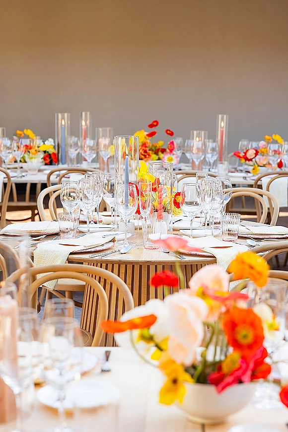 Reception tablescape with wedding reception table setting on a round wooden table, colorful floral centerpieces, glass cylinder candles, and bentwood chairs