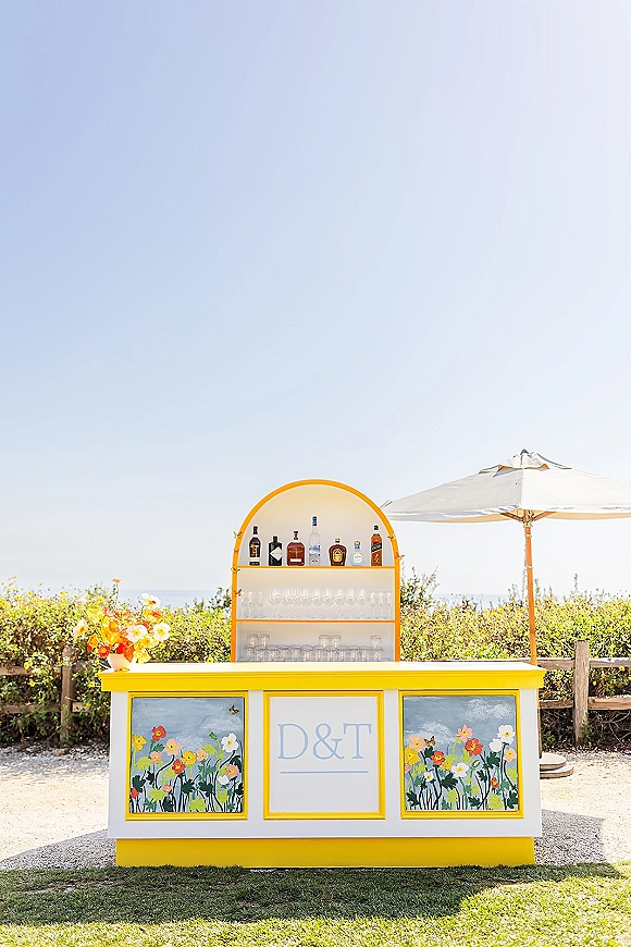Wedding bar setup with an outdoor wedding bar cart, liquor bottles and glassware, plus a monogram sign overlooking the ocean under a patio umbrella