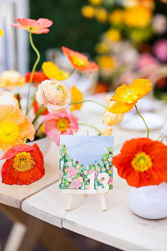 Wedding table number card on a small easel showing 13, framed by poppy and ranunculus centerpieces on a wood reception table