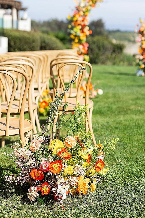 Ceremony aisle decor with colorful aisle flowers lining bentwood chairs, leading to a floral arch on a grass lawn with hedges and trees
