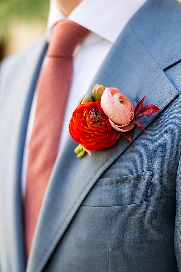 Groom boutonniere with ranunculus boutonniere on a blue suit jacket and coral tie, greenery in lapel buttonhole, soft garden bokeh behind