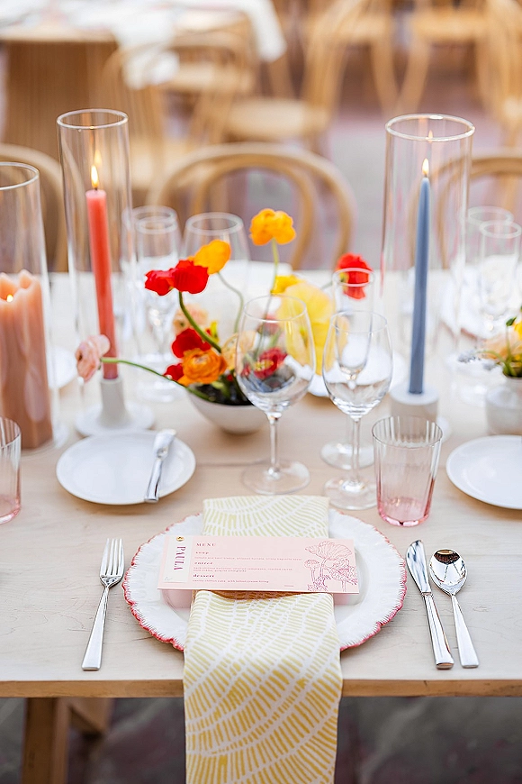 Reception tablescape with wedding place setting, taper candles in glass hurricanes, floral centerpiece, menu card on plates, and wood chairs in back