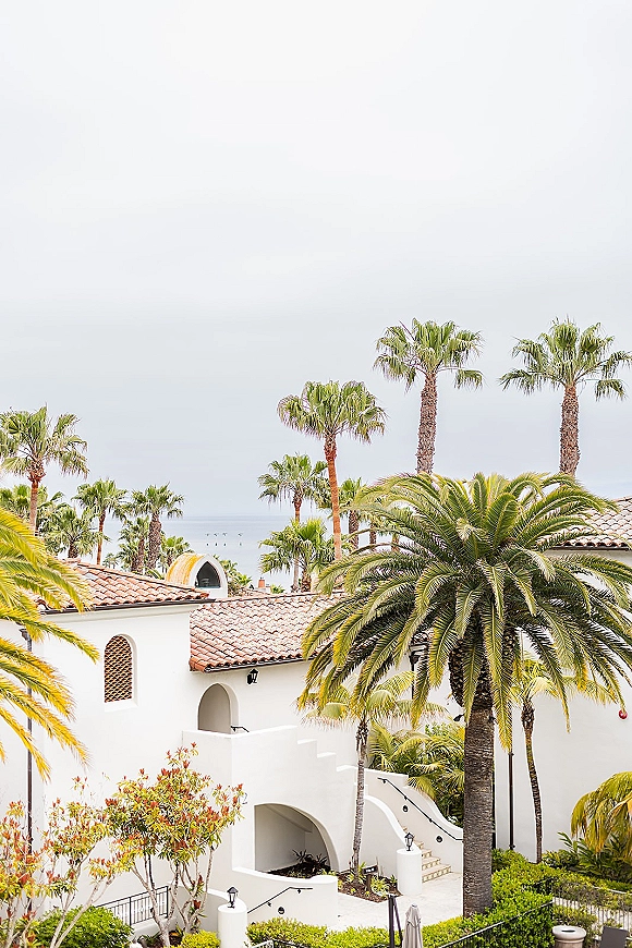 Wedding venue exterior with white stucco walls, arched windows, and palm trees, featuring an outdoor stairway and ocean view beyond