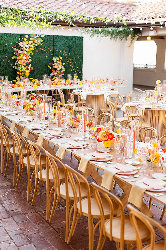 Reception tablescape with long banquet table setting on an outdoor patio, white linens, orange-pink florals, candles, and string lights overhead