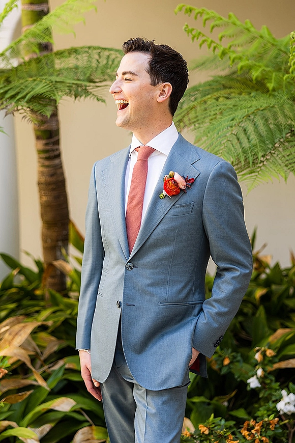 Groom portrait in a light blue suit laughing and looking away, hand in pocket with boutonniere, amid tropical greenery by a beige wall