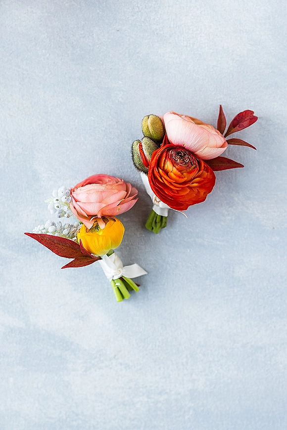 Wedding boutonniere with ranunculus bloom, greenery, and red foliage, wrapped in white ribbon, resting on a light blue surface