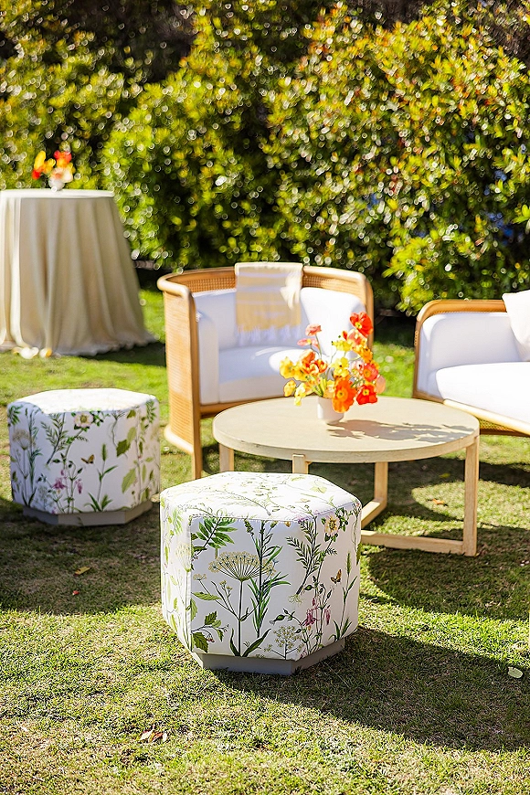 Lounge seating area with wedding lounge furniture, a white rattan sofa, round coffee table, floral ottomans, on a sunny garden lawn