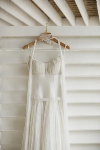 Wedding dress on hanger with tulle skirt and beaded straps, a sheer scarf wrap draped against a white wood wall with beams