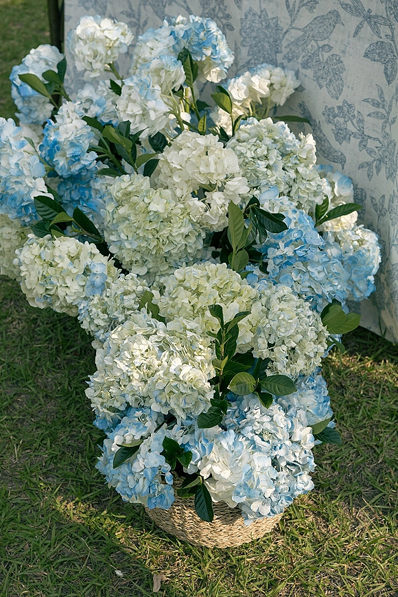 Hydrangea arrangement of blue and white hydrangeas in a woven basket with green leaves on a grass lawn beside a patterned chair