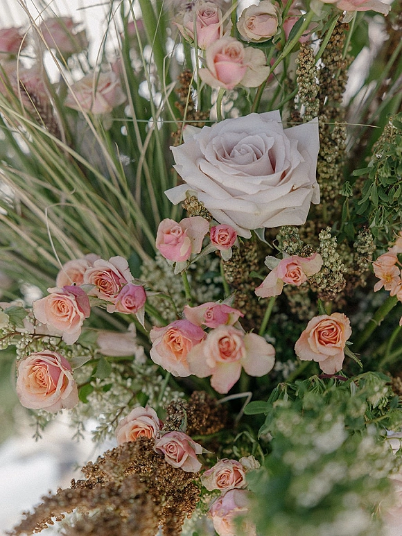 Wedding florals featuring rose wedding flowers with blush roses, greenery and tall grasses arranged outdoors against sky and foliage backdrop