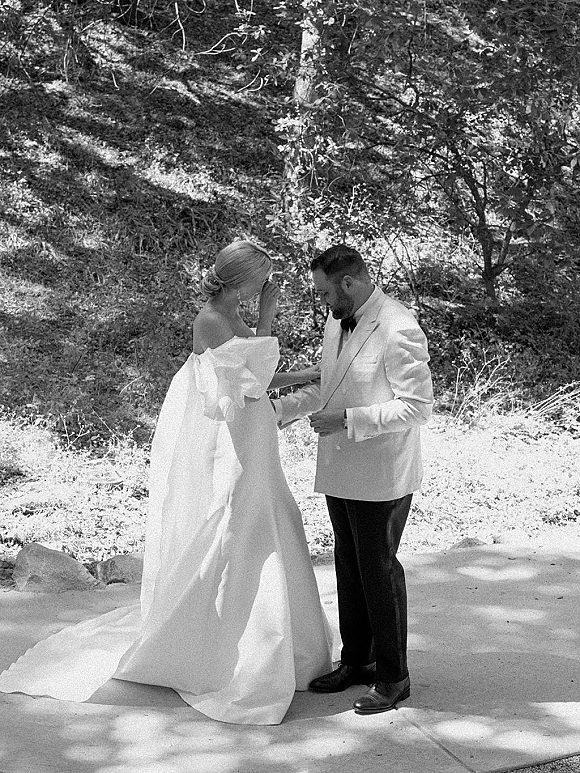 First look moment outdoors as bride wipes tears and holds the groom’s hands on a dappled woodland path, her strapless dress train flowing