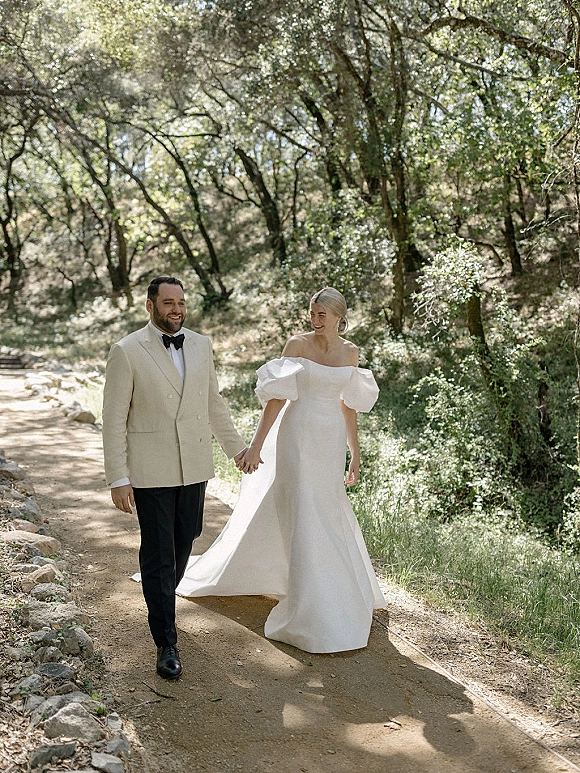 Couple portrait of bride and groom holding hands, walking a sunlit woodland path; her puff-sleeve off-shoulder gown trails behind.