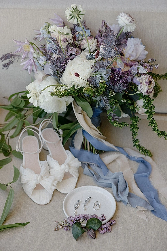Wedding details flat lay with bridal details flat lay elements—bouquet, bow heels, rings, pearl drop earrings and blue silk ribbon on linen backdrop