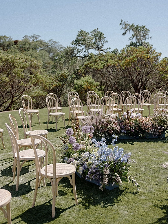 Ceremony setup with outdoor wedding ceremony seating, bentwood chairs flanking a meadow of blue and lavender aisle flowers on a lawn under blue sky