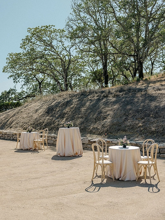 Outdoor cocktail tables set for wedding cocktail hour with ivory linens and small floral centerpieces on a hillside patio by a stone wall