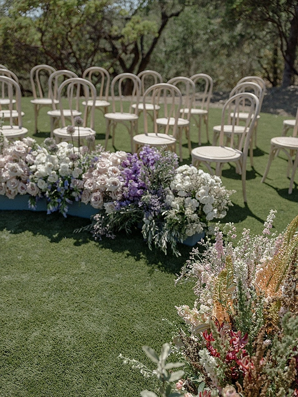 Ceremony aisle decor with outdoor ceremony chairs and low rose-and-wildflower arrangements lining a grassy garden aisle under trees