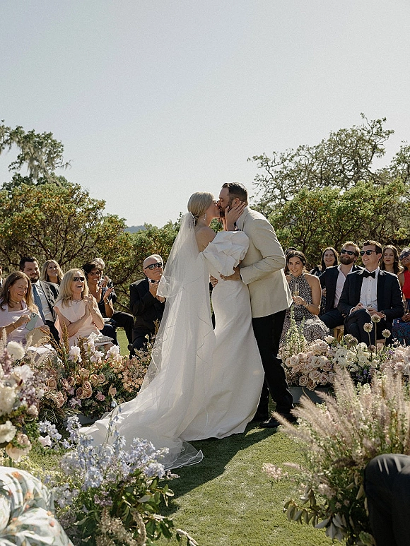 Wedding kiss as the bride holds the groom’s face, long veil flowing beside meadow aisle florals, guests seated in a sunny garden ceremony